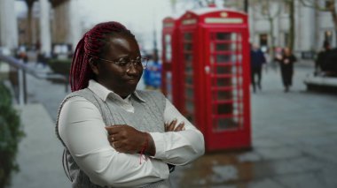 Şehrin arka planında kollarını kavuşturmuş kırmızı telefon kulübesi olan bir kadın açık şehir ortamını öneriyor..
