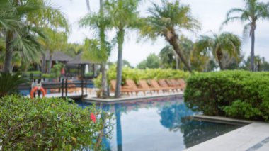 Palmiye ağaçları, yüzme havuzu ve yemyeşil bitkilerle çevrili uzanmış dinlenme odalarının bulunduğu lüks bir tropikal tatil köyünün bulanık manzarası dingin bir ortam yaratıyor..