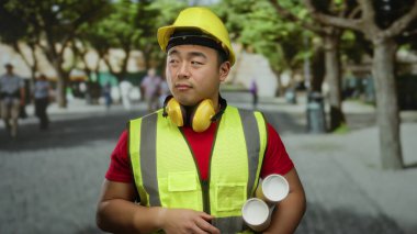 Kentsel caddede güvenlik ekipmanı giymiş genç bir adam elinde planlar, açık bir şehir ortamında mimari planlama ve inşaatı sembolize eden dostane bir jest sunuyor..
