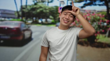 Genç adam, ağaçlar ve arabalarla çevrili güneşli bir caddede el hareketi yapıyor, kendine güven ve neşe saçıyor..
