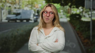 Gözlüklü genç sarışın kadın, kalabalık bir şehir caddesinde güvenli bir şekilde kollarını kavuşturarak şehir hayatı ve sakin bir açık hava ortamının bir karışımını yakalıyor..