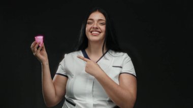 Pembe bir adet bardağını tutarken gülümseyen ve işaret eden bir kadın izole edilmiş siyah bir arka plana karşı, çevre dostu kadın bakımı çözümlerini güvenle destekliyor..