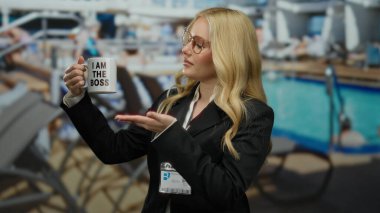 Gemi güvertesinde patron kupasını tutan kadın, havuz kenarında boş bir arkaplan karşısında liderlik ve çalışma ortamında kendine güvenini gösteriyor..