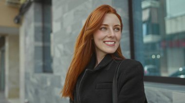 Şehirli bir caddede, uzun kızıl saçlı ve siyah ceketli bir kadın güvenle gülümsüyor. Şehir zeminine karşı. Modern bir yaşam tarzıyla güzellik ve moda sergiliyor..