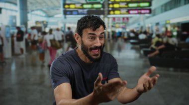 İspanyol asıllı, sakallı bir adam yoğun havaalanı terminalinde arka planda bulanık yolcular ve tabelalarla açıkça işaret veriyor..