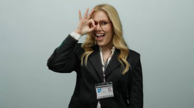 Gözlüklü kadın, gri arkaplan karşısında el hareketi yapıyor. İş elbisesi kimliğiyle gülümsüyor. Kendine güvenen sarı saçlı..