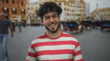 Sakallı, gözlüklü, açık havada gülümseyen kırmızı çizgili tişörtlü ve arka planda insanlar olan İspanyol bir adam..