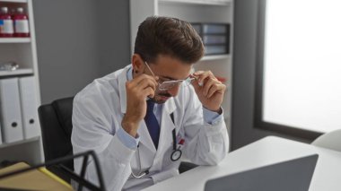 Hastane odasında beyaz önlük giyen genç bir adam masasında otururken gözlüklerini ayarlıyor, klinik ortamda odaklanmış ve profesyonel bir atmosfer yansıtıyor..