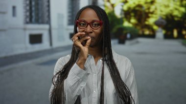 Şehir ortamında kırmızı gözlüklü, samimi bir gülümseme takan ve sessiz bir jest yapan kadın, örgü örüyor ve sokak ortasında günlük tarzını belirtiyor.