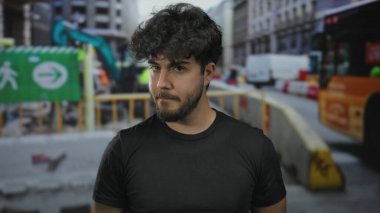 Sakallı, genç İspanyol bir adam. Yoğun bir inşaat sahasında duruyor. Çeşitli ekipman ve yapılarla..