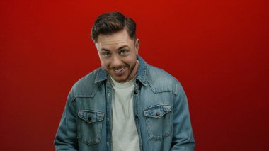 Kot gömlekli sakallı genç adam canlı kırmızı arka planda dalgın dalgın bakıyor.