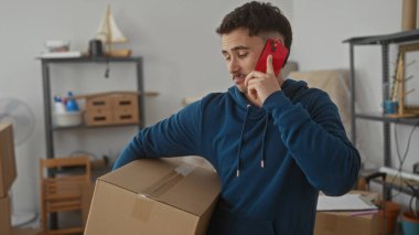 Genç adam yeni bir apartman dairesinde karton bir kutu tutarken akıllı telefondan konuşuyor. Etrafı eşyalarla çevrili, hareket sürecini sembolize ediyor..