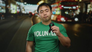 Yeşil tişörtlü genç gönüllü adam, arka planda bulanık ışıklar, farkındalığı ve hatıraları simgeleyen hareketli bir şehir caddesinde gece vakti siyah kurdele tutuyor..