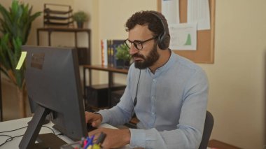 Ofiste çalışan, kulaklık takan, bilgisayar kullanan, kapalı alandaki bitkiler ve profesyonel çevreyi geliştiren organize raflar kullanan İspanyol bir adam..