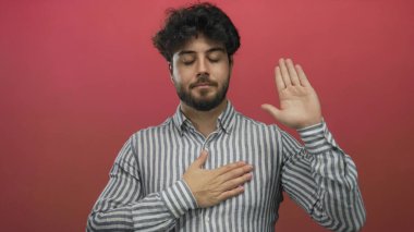 Sakallı, kırmızı arka planda yemin jesti yapan, çizgili tişörtlü genç İspanyol bir adam..