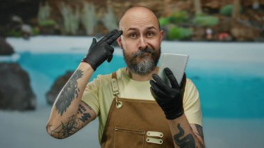 Sakallı ve eldiven takan bir adam defterini otelin açık havuzunda, yemyeşil bir otelin ortasında tutuyor..