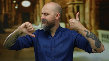 Mavi gömlekli kel bir adam kapalı bir kilisede parmak uçlarında çelişkili duygular ve dövmeler sergiliyor bulanık bir arkaplanda..