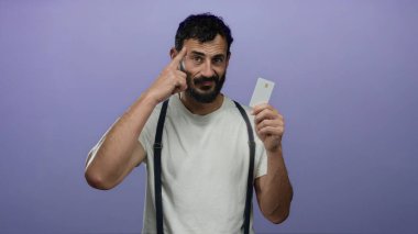 Mor arka planda kredi kartı tutan İspanyol bir adam, beyaz gömlek ve pantolon askısı giyiyor, kafasına doğru hareketler yapıyor, düşünceli finansal kararları öneriyor..