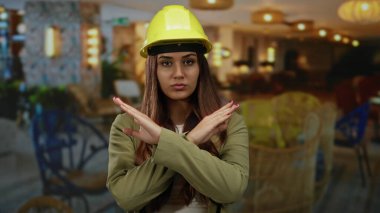 Sarı kasketli kadın mimar, şık otel odasında X hareketi yapıyor ve güçlü profesyonel duruşuyla reddediyor.