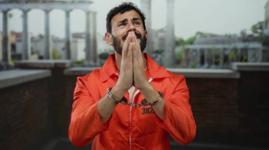 Turuncu tulumlu, sakallı ve kelepçeli İspanyol bir adam eski Roma düzeninde antik mimari arka planda el ele tutuşup yalvarıyor..