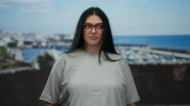 Gözlüklü kadın deniz kenarındaki limanın yanında duruyor. Kıvrımlı moda ve plaj sahnesini kucaklıyor. Rahat bir tavırla..