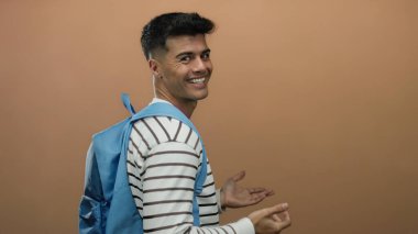 Sırt çantalı genç adam gülümsüyor ve izole bir kahverengi duvara başparmak işareti veriyor. Neşeli ve kendinden emin bir tavır sergiliyor..