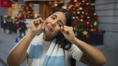 Şehir sokağında açık bir kadın, süsleri şenlikli. Soğuk bir günde arka planda ışıklar yanıp sönerken, yılbaşı ağacının yanında günlük beyaz giysiler içinde üzgün görünüyor..