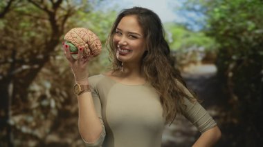 Parkta elinde saatli bir beyin mankeni olan bir kadın, İspanyol kökenli, bej giyen, açık hava ortamında yemyeşil alanlar arasında düşünceli bir şekilde gezinen..