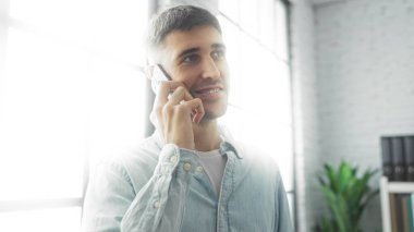 Geniş pencereli, parlak, modern ofis içinde telefonla konuşan genç adam profesyonel ve sıradan bir atmosfer sergiliyor, odak ve iletişimi vurguluyor..