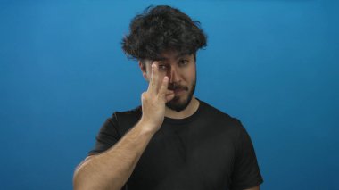 Sakallı genç adam mavi bir arka plana karşı jestler yapıyor. Hispanik özellikleri ve günlük kıyafetleri olan tarafsız bir ortamda iletişimi ve ifadeyi vurguluyor..