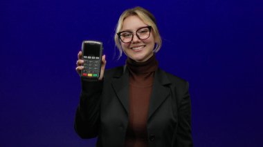 Ödeme terminalini mavi arka planda gösteren kadın, ekonomik işlemler için kullanım kolaylığı ve modern teknolojiyi neşeli bir ifade ve iş kıyafeti ile gösteriyor..