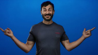 Günlük gömlekli İspanyol bir adam gülümsüyor ve parlak mavi bir arkaplanı işaret ediyor. Olumlu duygular ve gelişigüzel davranışlar sergiliyor..