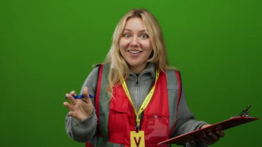 Sarı yelekli, yeşil yelekli, elinde not panosu ve yeşil arka plan duvarında kalem tutan genç bir gönüllü sürpriz ya da heyecan ifade ediyor..