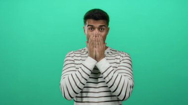 Genç adam elleri ile ağzını kapatıyor yeşil bir zemine karşı neşeli bir şekilde duruyor çizgili bir gömlek giyiyor duygularını ve şaşkınlığını gösteriyor..