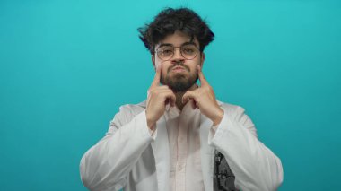 Laboratuvar önlüklü, gözlüklü ve sakallı genç adam mavi bir arka plana karşı duruyor. Profesyonel bir tıbbi ortamda İspanyol bir doktor ya da bilim adamını canlandırıyor..