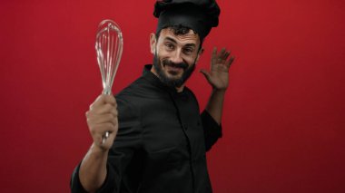 Sakallı İspanyol erkek aşçı neşeli bir şekilde çırpıcıyı canlı kırmızı arka planda tutuyor, aşçılık hevesini ve profesyonel kıyafetlerini sergiliyor..