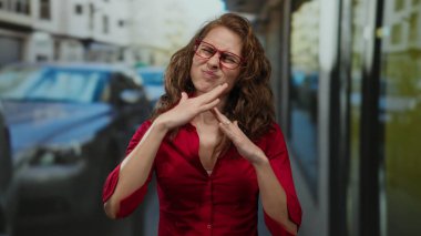 Kırmızı gözlüklü ve kıvırcık saçlı bir kadın şehir caddesinde el hareketi yapıyor, kırmızı bir gömlek giyiyor ve açık hava ortamında düşünceli görünüyor..