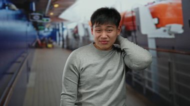 Şaşırmış bir ifadeye sahip genç bir adam, açık deniz manzarası ile çevrili bir yolcu teknesinde duruyor, canlı bir gemi teçhizatı ve yapılara karşı düşünceli bir görünüm sergiliyor.