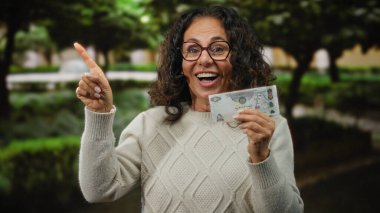 Parkta orta yaşlı bir kadın elinde sürpriz bir ifadeyle Dirham banknotunu tutuyor. Heyecan ya da keşif öneriyor..