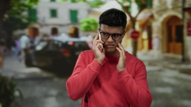 Gözlüklü ve kırmızı kazaklı İspanyol bir adam işlek bir şehir caddesinde akıllı telefondan konuşuyor, bulanık bir şehir geçmişiyle ilgili endişelerini aktarıyor ve hareketli atmosferi güçlendiriyor.