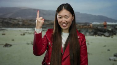 Sahilde kırmızı ceketli, arka planda dağlarla gülümseyen neşeli ifadeler sergileyen ve açık gökyüzünün altında işaret eden bir kadın..