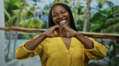 Sarı tişörtlü kıvırcık saçlı bir kadın havuz başında gülümserken elleriyle kalp şeklinde bir kalp yapıyor, açık havada neşeli bir tropikal titreşim yakalıyor..