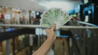 Adam alışveriş merkezinin içinde dolarları tutuyor, alışveriş ortamında banknotlar sergiliyor, finans, perakende ve kapalı alanlarda harcama temaları vurguluyor..