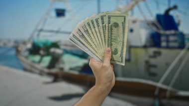 Arka planda tekneler, finans, seyahat ve deniz hayatı konseptini gözler önüne seren, açık hava ortamını ve Kafkas kökenini vurgulayan Amerikan dolarlarını sahil limanında tutan adam..