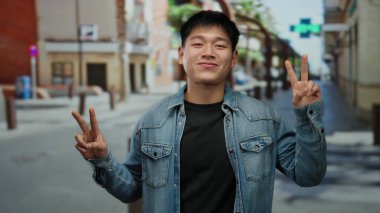 Şehir sokaklarında barış işareti gösteren, kot ceket giyen, arka planda şehir binaları olan, kaygısız yaşam tarzını, Asya kültürünü ve modern açık hava eğilimlerini temsil eden genç bir adam..