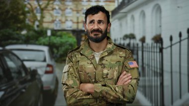 Asker üniforması giyen sakallı bir adam şehir sokaklarında arabalarla dikiliyor ve ciddi bir ifade sergiliyor..