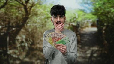 Parkta Norveç banknotları tutan bir adam, arka planda yeşillik ve güneş ışığı ile sürpriz ifade ediyor, günlük kıyafetler giyiyor ve çeşitli para birimlerini gösteriyor..