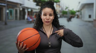 Sokakta basketbol oynarken kendini gösteren bir kadın, arka planda bulanık bir şehir ortamında kendine güvenen bir İspanyol kadın koçu gösteriyor..