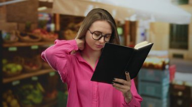 Meyve pazarında kitap okuyan, gözlüklü ve pembe gömlekli, arka planda çeşitli meyveler olan bir kadın, düşünceli bir sahne iletiyor.