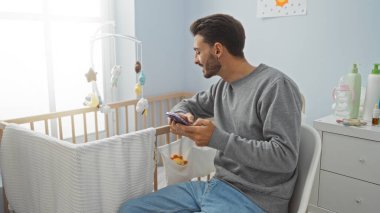 Bebek odasında beşikte oturan İspanyol bir adam, elinde akıllı bir telefon ve mutlu görünüyor, bebek malzemeleri, oyuncaklar ve hafif ışıklandırma ile çevrili, rahat bir evde..